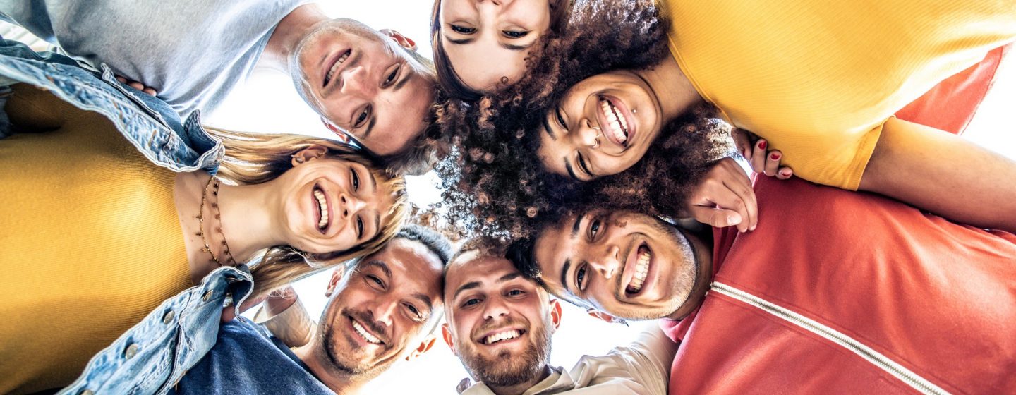 Multiracial group of young people standing in circle and smiling at camera - Happy diverse friends having fun hugging together - Low angle view